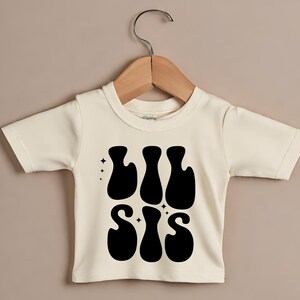 Op de afbeelding: Een witte baby romper met een zwarte afbeelding waarop "Lil Sis" staat in een retro lettertype.