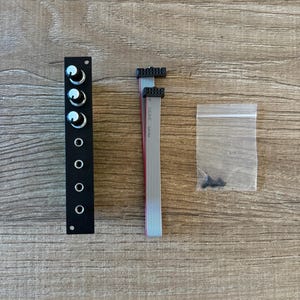 Könnte beinhalten: Eine schwarze Platte mit sechs silbernen Knöpfen, ein graues Flachbandkabel mit schwarzen Steckern und eine durchsichtige Plastiktüte mit schwarzen Schrauben.