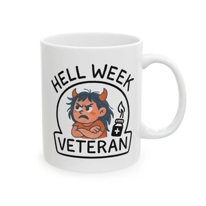 以下が含まれることがあります： 白のセラミックマグカップ。イラストは白黒で、腕を組んだ漫画の悪魔と「HELL WEEK VETERAN」の文字が描かれています。プラス記号の付いた小さなボトルも描かれています。