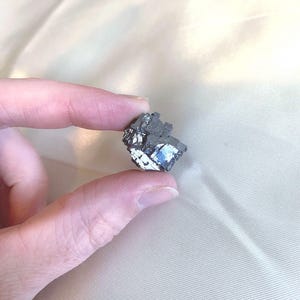 May include: A small, irregularly shaped, metallic mineral specimen. The mineral has a dark, textured surface with areas of silver and blue iridescence. It is held between two fingers against a neutral background.
