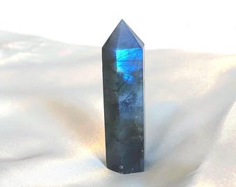 May include: A polished labradorite crystal point. The crystal is a dark gray color with flashes of blue and iridescent hues. The top of the crystal is a pointed shape. The crystal is displayed on a white, silky fabric.