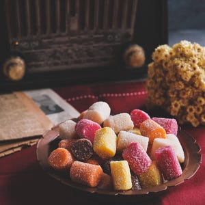 May include: A copper-colored plate filled with colorful, sugared candies. The candies are various shapes and colors, including pink, yellow, orange, and white. A vintage radio and dried flowers are in the background.