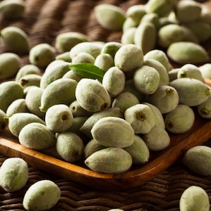 May include: A wooden bowl filled with fresh, green almonds. The almonds are oval-shaped and covered in a soft, fuzzy coating. A few green leaves are scattered among the almonds, and the bowl sits on a woven surface.