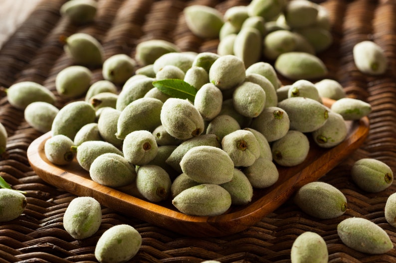 May include: A wooden bowl filled with fresh, green almonds. The almonds are oval-shaped and covered in a soft, fuzzy coating. A few green leaves are scattered among the almonds, and the bowl sits on a woven surface.