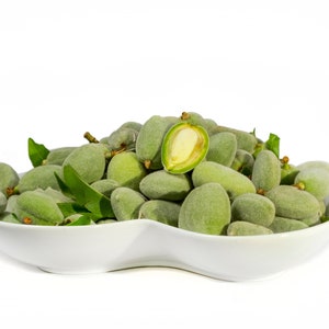 May include: A white, oval-shaped bowl filled with fresh, green almonds. One almond is cut open, revealing the inner nut. Green leaves are interspersed among the almonds. The background is plain white.