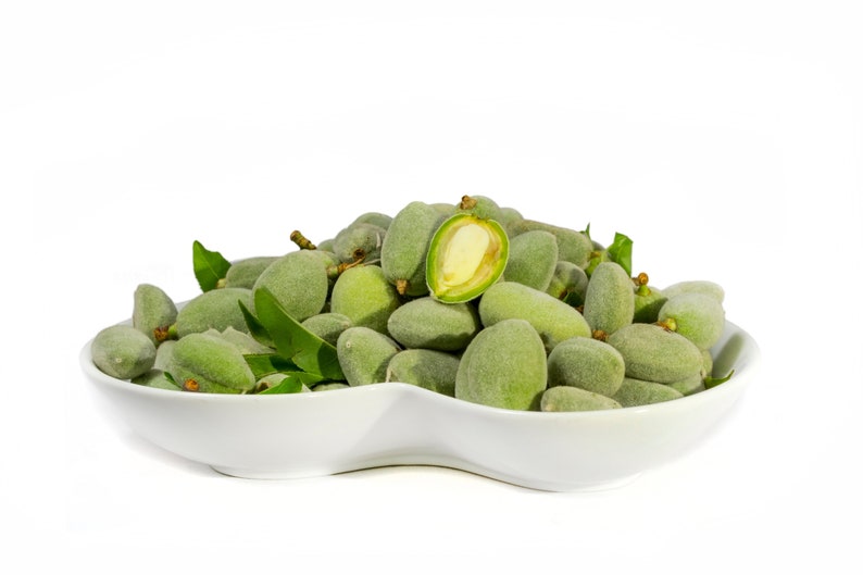 May include: A white, oval-shaped bowl filled with fresh, green almonds. One almond is cut open, revealing the inner nut. Green leaves are interspersed among the almonds. The background is plain white.