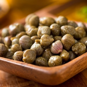 Puede incluir: Un bol de madera lleno de alcaparras verdes. Las alcaparras son pequeñas, redondas y tienen una textura ligeramente arrugada.