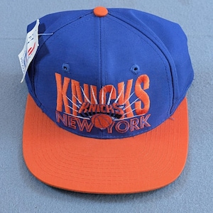 Puede incluir: Gorra de béisbol azul y naranja con el texto "Knicks New York" en naranja. La gorra tiene una visera plana y un botón en la parte superior. Tiene una etiqueta adjunta.