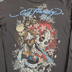 May include: Dark gray long-sleeve shirt featuring a skull graphic with floral and rhinestone accents. The shirt has the text "Ed Hardy" in light blue script across the top. The graphic includes red, pink, and white flowers.