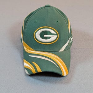 May include: Green and gold baseball cap featuring the letter "G" in a white oval with a gold outline. The cap has a curved brim with gold and white accents. A small button is on top.