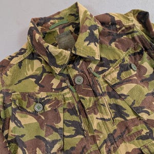 May include: Camouflage jacket in shades of green, brown, and black. The jacket features a collared neckline, two chest pockets with button closures, and a front zipper. The fabric has a textured appearance.