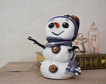 May include: A whimsical snowman figurine with a white body, black twig arms, and a carrot nose. It wears a blue and purple scarf and hat, and has brown button details. The snowman is a decorative winter holiday item.