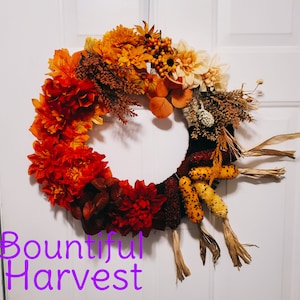 Può includere: Una ghirlanda autunnale con fiori arancioni, rossi e gialli, foglie marroni e pannocchie di mais. La ghirlanda è decorata con il testo "Bountiful Harvest".