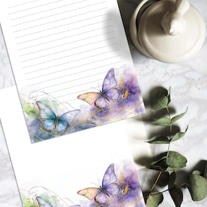 Może przedstawiać: Biały papier listowy w linie z akwarelowymi wzorami motyli w odcieniach niebieskiego, fioletowego i żółtego. Papier listowy posiada ozdobną obwódkę ze złotymi akcentami. Widoczny jest również mały, biały ceramiczny pojemnik z pokrywką w kształcie ptaka.
