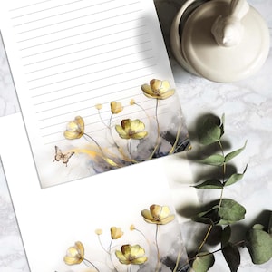 Może przedstawiać: Dwa arkusze papieru listowego w linie z akwarelowym wzorem żółtych kwiatów i motyli. Kwiaty są jasnożółte z złotym połyskiem. Motyle są brązowe z jasnobrązowym wzorem.