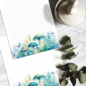 Może przedstawiać: Dwa arkusze papieru w linie z akwarelową ilustracją niebieskich grzybów i motyli na dole każdego arkusza. Grzyby otoczone są zielonymi liśćmi i kwiatami.