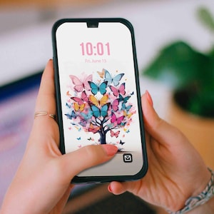 Könnte beinhalten: Ein schwarzes Smartphone, das ein farbenfrohes Schmetterlingsbaum-Design auf seinem Bildschirm zeigt. Der Bildschirm zeigt die Uhrzeit 10:01 Uhr und das Datum Freitag, 15. Juni. Das Telefon wird von einer Person mit Ring und Armband gehalten.