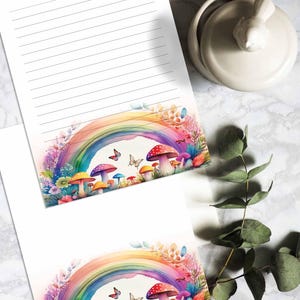 Może przedstawiać: Dwa białe arkusze papieru z nadrukowanym na dolnej krawędzi tęczą, grzybami i motylami. Tęcza jest wielokolorowa, a grzyby są czerwone, żółte i pomarańczowe. Motyle są fioletowe i niebieskie.