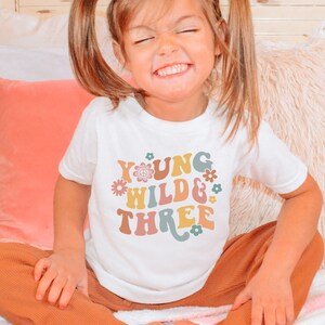 May include: A white t-shirt with a colorful graphic that says "Young Wild & Three" with flowers and a peace sign. The shirt is being worn by a young child with blonde hair.