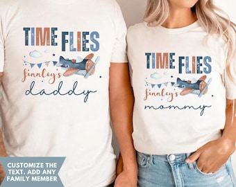 May include: Two white t-shirts with blue and orange text that reads "Time Flies" and a small airplane graphic. The text below the airplane says "Finley's" and "daddy" on one shirt and "mommy" on the other.