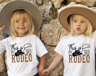 Könnte beinhalten: Zwei Kinder tragen weiße T-Shirts mit dem Text "This is my 3rd Rodeo" und einer Cowboyhut-Grafik. Die Kinder halten sich an den Händen und tragen Cowboyhüte.