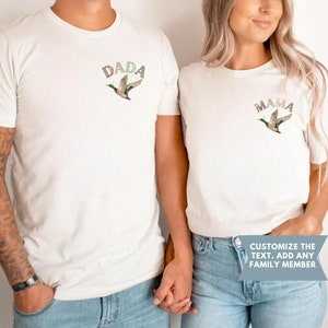 May include: Two white t-shirts with a flying duck and the words "DADA" and "MAMA" printed on them. The text is in a brown, pink, and green colour scheme. The shirts are being held by two people.