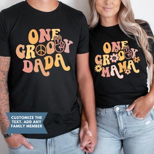 Może przedstawiać: Dwie czarne koszulki z retro wzorem z lat 70. Tekst na koszulkach brzmi "One Groovy Dada" i "One Groovy Mama".
