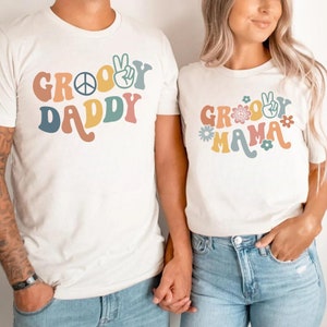 May include: Two white t-shirts with a retro groovy font design. The shirt on the left says "Groovy Daddy" with a peace sign and a peace hand gesture. The shirt on the right says "Groovy Mama" with flowers and a peace hand gesture.