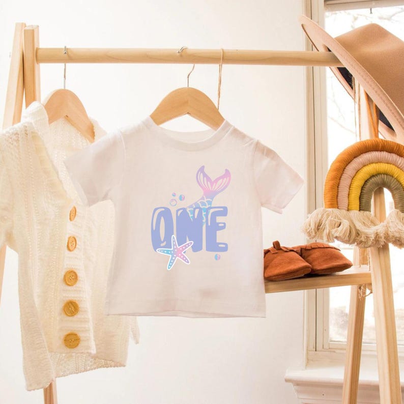May include: White baby onesie with a blue and pink mermaid tail and the word "ONE" in bubble letters. A starfish is below the word "ONE".