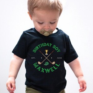 May include: Black t-shirt with a green graphic of crossed golf clubs, a golf ball, a golf hat, and a trophy. The text "Birthday Boy" is above the graphic and "EST. 2023" and "MAXWELL" are below the graphic.