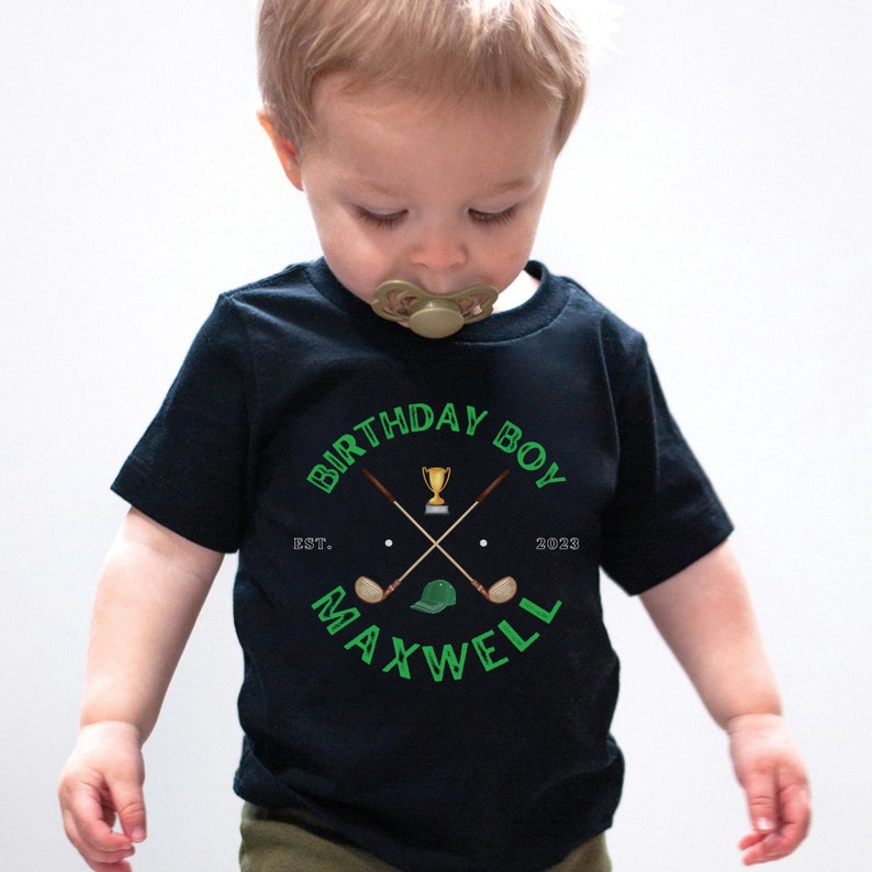 May include: Black t-shirt with a green graphic of crossed golf clubs, a golf ball, a golf hat, and a trophy. The text "Birthday Boy" is above the graphic and "EST. 2023" and "MAXWELL" are below the graphic.