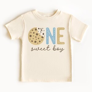 May include: Cream-colored toddler t-shirt with a cookie graphic and the words "ONE sweet boy" in blue, tan, and black. The cookie has a bite taken out of it, revealing chocolate chips. A cute and playful design for a first birthday.
