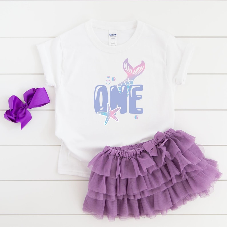 May include: A white t-shirt with a purple, blue, and pink mermaid tail and the word "ONE" in a bubble font. The shirt is paired with a purple tulle skirt.