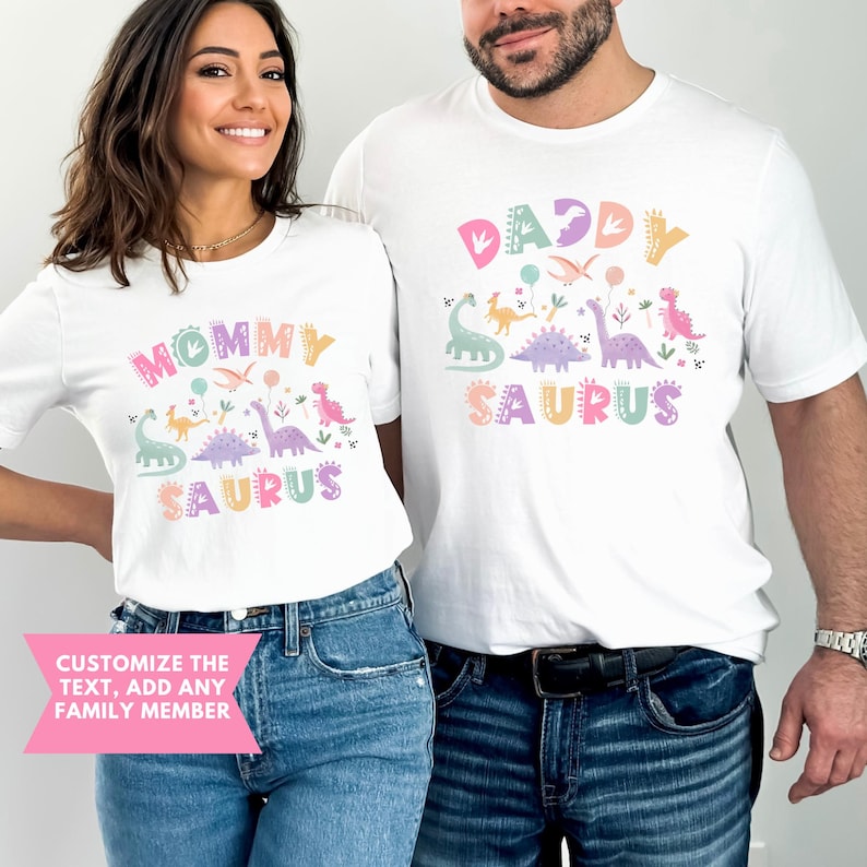 May include: Two white t-shirts featuring dinosaur illustrations and the phrases "Mommy Saurus" and "Daddy Saurus" in pastel colours. The image shows a couple wearing the shirts, suggesting a family-oriented product or gift idea.