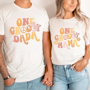 Może przedstawiać: Dwie białe koszulki z retro groovy wzorem czcionki. Tekst na pierwszej koszulce brzmi "One Groovy Dada", a tekst na drugiej koszulce brzmi "One Groovy Mama".