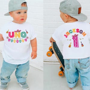 May include: White t-shirt with the word "UNO" in colorful letters, a sombrero, and guitars. The back of the shirt says "JACKSON" and the number "1" with a sombrero and guitars. The child is wearing blue jeans and a gray cap.