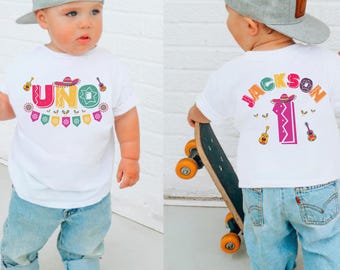 May include: White t-shirt with the word "UNO" in colorful letters, a sombrero, and guitars. The back of the shirt says "JACKSON" and the number "1" with a sombrero and guitars. The child is wearing blue jeans and a gray cap.