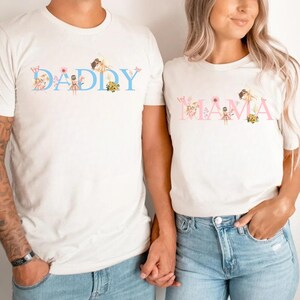 May include: Two white short-sleeve t-shirts. The left shirt features the word DADDY in light blue with fairy and floral illustrations. The right shirt features the word MAMA in light pink with fairy and floral illustrations. Both have crew necks.