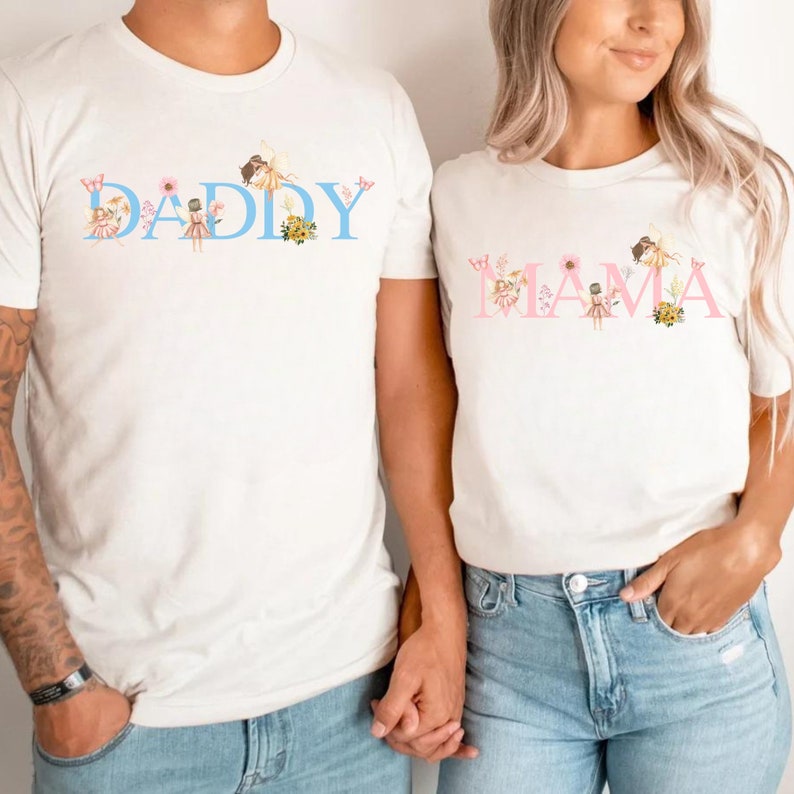 May include: Two white short-sleeve t-shirts. The left shirt features the word DADDY in light blue with fairy and floral illustrations. The right shirt features the word MAMA in light pink with fairy and floral illustrations. Both have crew necks.