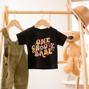 Może przedstawiać: Czarny T-shirt z kr&oacute;tkim rękawem z kolorowym nadrukiem, na kt&oacute;rym widnieje napis "ONE GROOVY BABE" wraz ze znakiem pokoju i kwiatami.