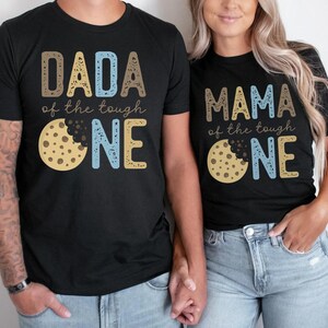 May include: Black t-shirts with matching designs. One shirt says "DADA of the tough ONE" with a cookie graphic. The other says "MAMA of the tough ONE" with a cookie graphic. The text and cookie are in shades of brown, blue, and yellow.