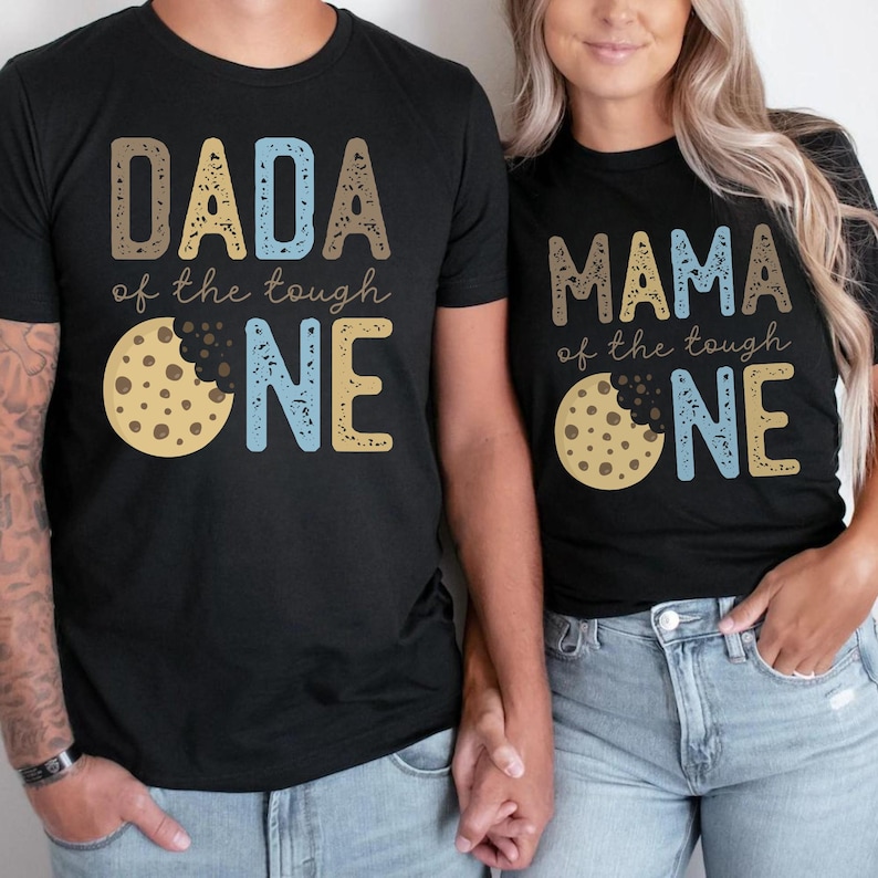 May include: Black t-shirts with matching designs. One shirt says "DADA of the tough ONE" with a cookie graphic. The other says "MAMA of the tough ONE" with a cookie graphic. The text and cookie are in shades of brown, blue, and yellow.
