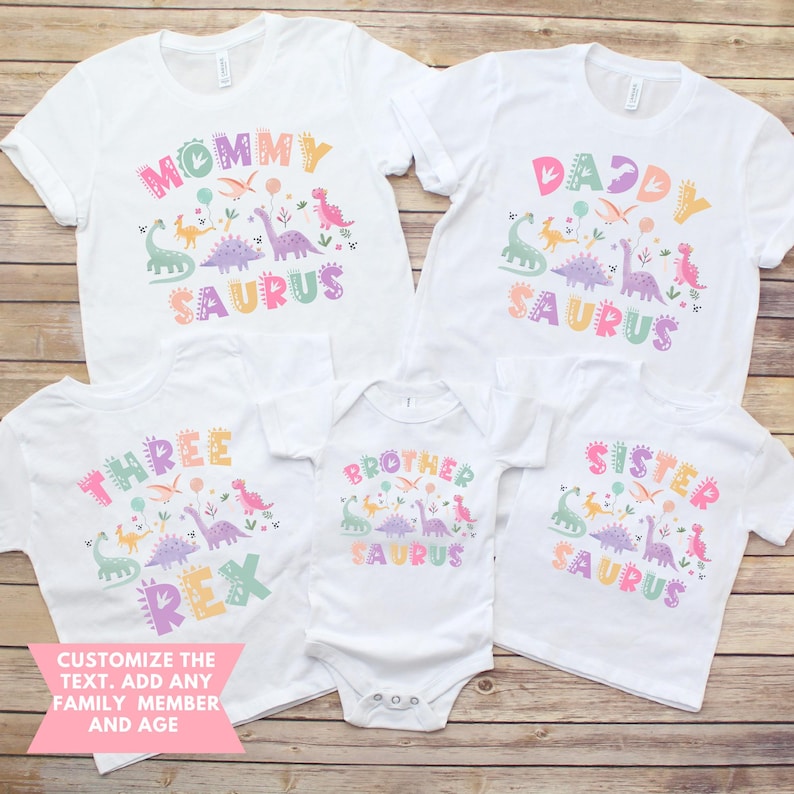 May include: A collection of white t-shirts and a babygrow, each featuring a dinosaur design and text. The shirts display phrases like "Mommy Saurus", "Daddy Saurus", "Three Rex", "Brother Saurus", and "Sister Saurus". Customisation is offered.