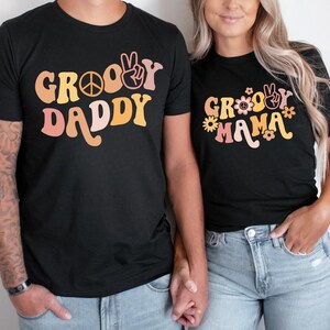 May include: A couple wearing matching black t-shirts with a groovy font design. The man's shirt says "Groovy Daddy" and the woman's shirt says "Groovy Mama".