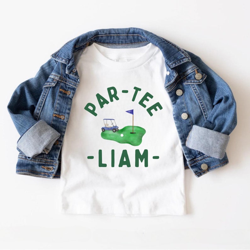 May include: White t-shirt with a green golf-themed graphic and the text "PAR-TEE - LIAM". The graphic includes a golf cart, a golf ball, and a flag on a green. A blue denim jacket is draped over the shirt.