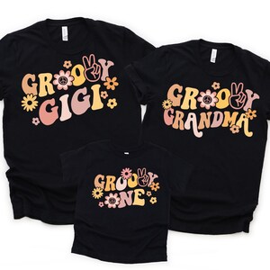 May include: Three black t-shirts with a groovy font design. The shirts say "Groovy Gigi", "Groovy Grandma", and "Groovy One". The shirts have a retro 70s vibe with peace signs and flowers.