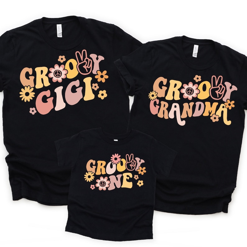 May include: Three black t-shirts with a groovy font design. The shirts say "Groovy Gigi", "Groovy Grandma", and "Groovy One". The shirts have a retro 70s vibe with peace signs and flowers.