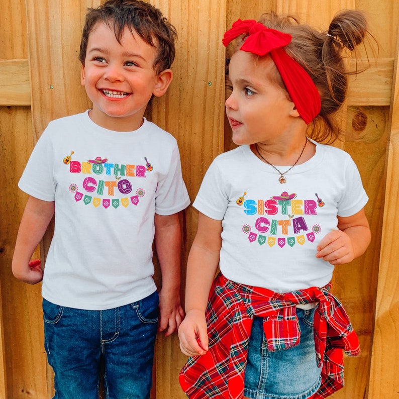 K&ouml;nnte beinhalten: Zwei Kinder tragen wei&szlig;e T-Shirts mit buntem Text, der "Brother Cito" und "Sister Cita" lautet. Die Shirts haben ein festliches, buntes Design mit einem Sombrero, einer Gitarre und Flaggen. Der Junge tr&auml;gt eine blaue Jeans und das M&auml;dchen einen rot-schwarz karierten Rock.