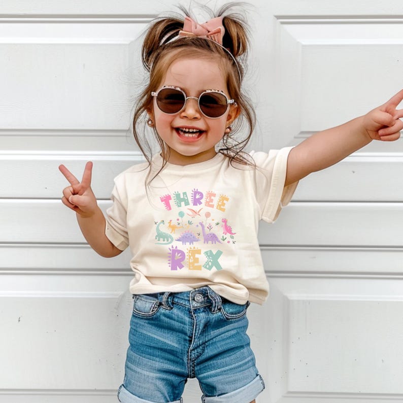 May include: A cream-coloured t-shirt with the text "THREE REX" in pastel colours, featuring dinosaur illustrations. The shirt is paired with denim shorts. The child is wearing sunglasses and a pink bow.