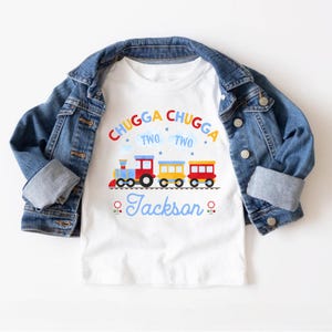 May include: White t-shirt with a colorful train graphic and the text "Chugga Chugga Two Two Jackson". The shirt is overlaid with a blue denim jacket. The train is red, yellow, and blue.